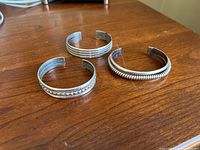 Three sterling silver cuff bracelets displayed on a wooden surface, showing overall condition and design details.