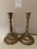 Front view of both silver candlesticks on flat surface against wall with handwritten tag above.