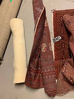 Three oriental woven rugs, two with red tones and intricate traditional patterns, one lighter colored rolled rug about 2.5 ft wide. Rugs are displayed unfolded and rolled on a concrete floor.