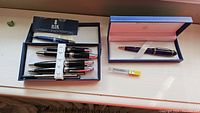 One Waterman pen in original blue box, four Bill Blass pens, Bill Blass packaging booklet, and yellow rollerball refill on table.