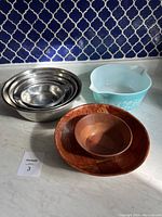 Photo showing all bowls in the lot: 5 stainless steel nested bowls, 2 wooden bowls (one larger carved, one smaller smooth), and one blue Pyrex glass bowl.