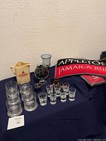 Various glassware including shot glasses, ceramic pitcher, and bar towels laid out on a dark cloth background