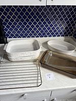 Photo showing all items together on countertop: two white porcelain dishes (one rectangular with floral, one oval), glass baking dishes stacked, metal oven rack in front