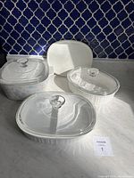 Four CorningWare ceramic dishes displayed including various shapes and sizes, with three glass lids and one plastic lid, on a white surface against a patterned blue tile background.