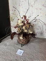 Overall view of dried flower arrangement in metal base planter on carpeted floor with floral wallpaper background.
