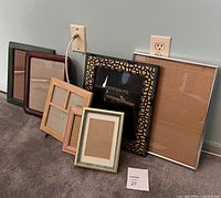 Six assorted picture frames resting against a wall, mostly wood frames in various sizes and one large silver metal frame