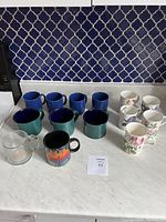 Photos of assorted mugs and fine bone china teacups on marble counter against navy patterned tile background showing quantity and styles.