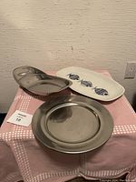 Front view of three serving dishes on a pink checkered cloth: Villeroy & Boch ceramic dish with fish design, oval divided stainless steel dish, and round stainless steel platter.