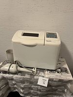 Black & Decker bread maker with digital control buttons and display on top, placed on table along with Braun hand blender and its cup, next to bread maker manual.