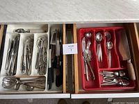 Drawer with multiple compartments filled with stainless steel cutlery and kitchen utensils, organized in white and red trays.