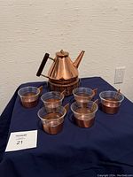 Full view of the entire copper tea set with teapot on burner and six cups with spoons around it on blue cloth