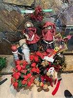 Photo showing two large cloth dolls in red and black dresses, artificial poinsettia flowers, plush reindeer, a colorful wooden toy, and assorted Christmas decorations grouped together on floor in front of a stone wall.