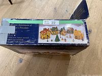 Original boxed packaging showing front of the Dickens Christmas Village set, depicting the complete village including houses, a Christmas tree, and figurines on the snow-covered base.