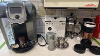 Full setup of Keurig coffee maker with accessories including reusable pod, glass carafe, metal cup with saucer, and travel mugs on kitchen counter.