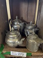 Four assorted silver and nickel plated tea and coffee pots arranged inside wooden cabinet shelf, showing variation in style and design with visible tarnish.