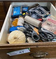 Open drawer showing assorted household tools and supplies such as staple gun, scissors, boxes of staples, string, tape measure.