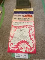 A collection of assorted sheet music displayed on the floor including Beautiful Brown Eyes, Whispering Hope, Whatever Will Be Will Be, Mockin Bird Hill, Puff The Magic Dragon, with a hardcover music book in a dark brown cover.