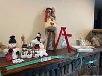 Wide shot of assorted snowmen and angel figurines displayed on a shelf, including fabric snowmen, ceramic/resin snowmen, and boxed angel ornaments.