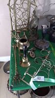 Photo of vintage brass and silver decor items on a green table including candle holders, goblet, small brass bell, brass fireplace tools, and a silver decorative item.
