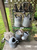 Full lot of vintage metal watering cans, toasters, metal bucket, rack and concrete sun decoration shown outside on wooden deck under wooden lattice.