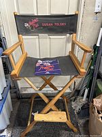 Front view of wooden director's chair with black fabric seat and embroidered backrest including 'Madame's Place' logo and 'Susan Tolsky'. Press kit placed on seat.