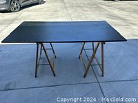 Two wooden collapsible sawhorse bases supporting a large black particle board tabletop outdoors on concrete.