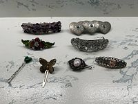 Photo shows full lot of nine hair accessories on white marbled background including large amethyst stone barrette, silver tone heart barrette with turquoise, sparkling faux diamond barrette, and various smaller clips and pins.