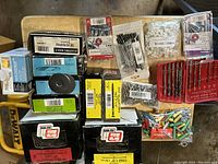 Various boxes and packages of screws, nails, plastic wall anchors, drill bits, and fasteners arranged on wooden surface. Boxes are dusty and unopened or partially unopened.