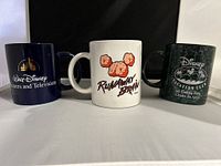 Front view of three Disney mugs: Walt Disney Pictures and Television mug in navy blue with gold logo; white Runaway Brain mug with evil Mickey Mouse brain silhouette; dark green Disney Vacation Club 4th birthday commemorative mug.