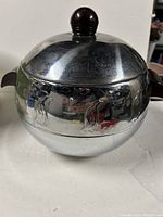 Close-up photo of the West Bend Penguin Hot Cold Server showing its spherical chrome body, black lid knob, and penguin engravings.