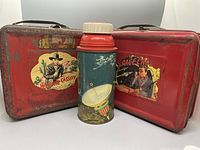 Two vintage metal lunchboxes and one metal thermos together showing wear and graphics