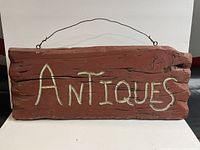 Front view of a wooden carved and painted 'ANTIQUES' sign with rustic folk art style and attached wire hanger.