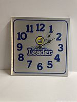 Front view of the square 1980s Leader farm supply clock showing the clock face with blue numbers and logo on brushed metal background.