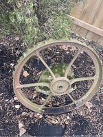 Top angle view of antique cast iron wheel showing six spokes and central hub; weathered with moss and rust; outdoors on soil near wooden fence