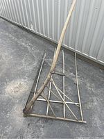 Full view of antique grain cradle scythe showing wooden handle and attached wooden cradle with five curved fingers lying on ground next to metal wall.