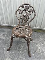 Frontal angled view of cast metal garden chair with rose floral pattern on backrest and circular floral cutout seat on asphalt beside metal siding.