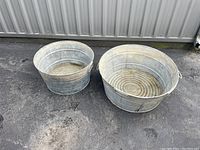 Top-side view of two vintage galvanized metal wash tubs placed on asphalt ground against metal wall, showing size difference and weathered surfaces.