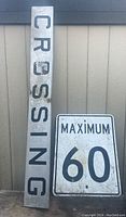 Photo showing both signs, vertical 'CROSSING' and square 'MAXIMUM 60', leaning against wall.
