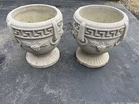 Pair of round concrete planters with Greek key pattern around top rim and neoclassical ornamental face motif on body, fluted pedestal base, placed outdoors on asphalt surface.