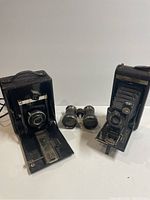 Two vintage folding cameras and one pair of binoculars shown together, cameras open with bellows extended.