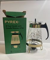 Photo of vintage Pyrex 8 cup coffee carafe sitting on gold-tone candle warmer stand next to green original box with product illustration and name.