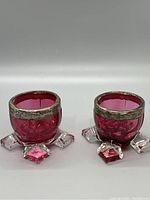 Pair of small cranberry glass egg cups with clear glass feet and heavily worn sterling silver rims shown front and angled view.