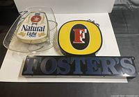 Three vintage advertising signs for beer brands including Natural Light, Foster's, and a yellow/black circular sign with a red F logo, shown against a white background.