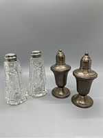 Photo showing four shakers: two clear cut crystal shakers with silver tops on left, two aged sterling silver weighted shakers on right.