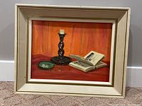 Framed oil on board painting showing a twisted black candle holder with a white candle, open book, and small green ashtray on a vibrant red fabric background, resting against a cream painted wooden frame.