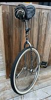 Full side view of the vintage unicycle showing the black seat with attached pouch, single white wall tire, pedals and chrome frame.