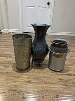 Photo shows all three decorative metal flower pots of varying sizes and styles placed on wooden floor in front of white door.