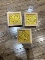 Three square wooden frames with yellow artificial rose flowers arranged inside, taken from a top angle on a wooden surface.