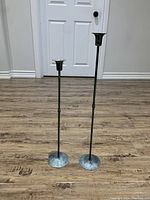 Two black metal candle holders of different heights placed on wooden floor with white door background