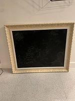 Chalkboard with ornate cream frame standing on floor against wall, showing some chalk markings on surface.